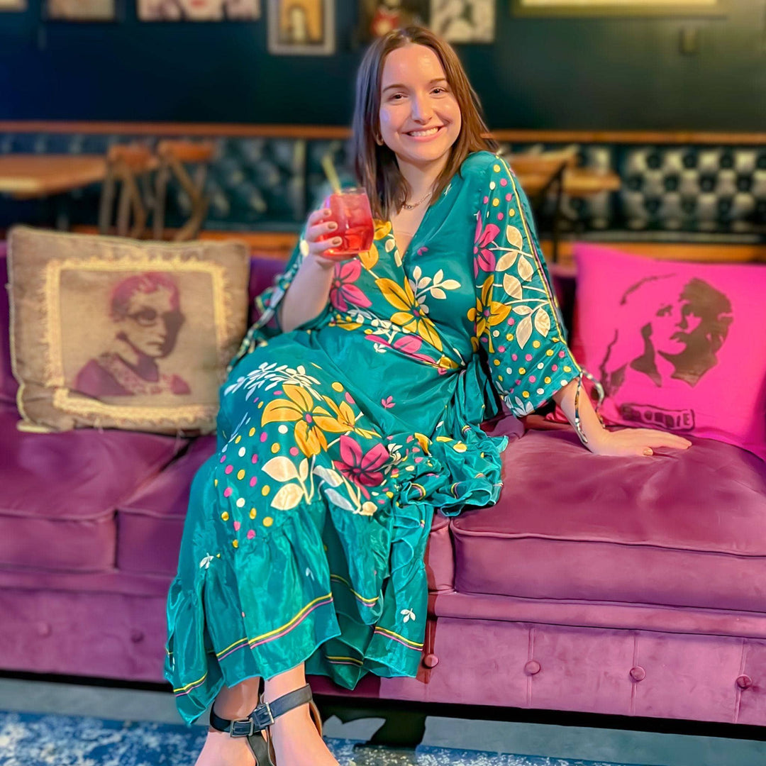 a woman sitting on a lavendar colored couch wearing a team zaria wrap dress. The dress has big white, pink and yellow flowers on it.
