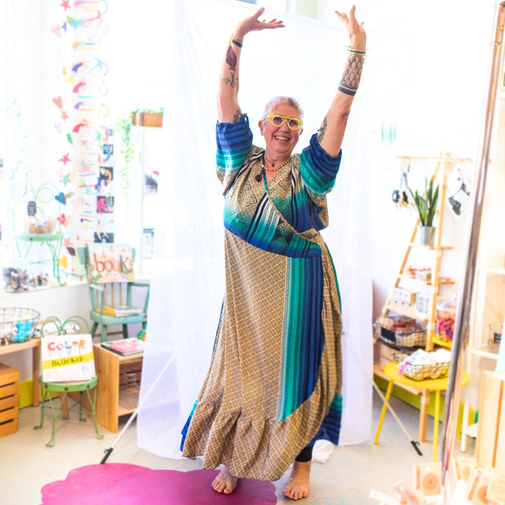 A woman barefoot dancing in a shop wearing a sari wrap dress. The majority of the dress is cream colored with blue ombre patterns along the sleeves and side of the dress.