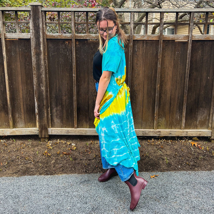 Woman in blue and yellow tie dye wrap dress worn as a duster, showing it off from the side while standing in front of a wooden fence.