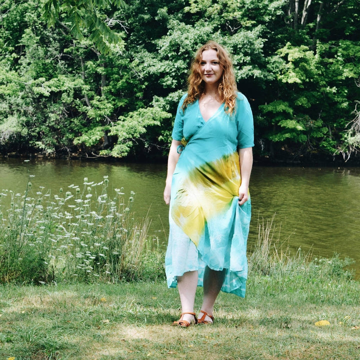 Model is walking outside in front of a stream wearing a tie-dye wrap dress in yellow and light blue.