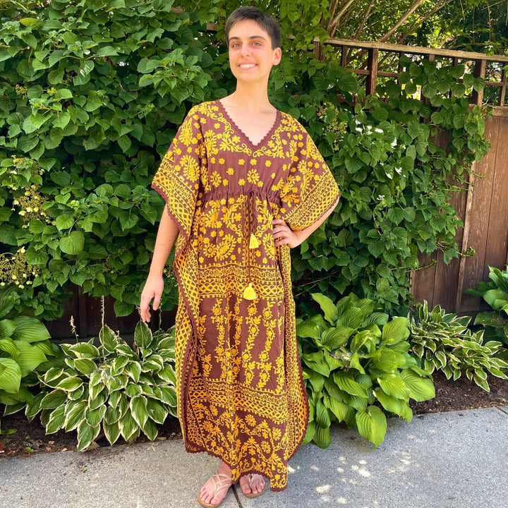 Model is wearing a brown/ yellow colorful embroidered kaftan while standing outside in front of a wall of greenery.