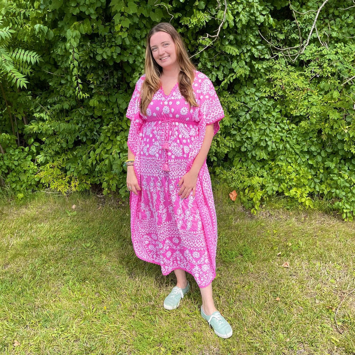 Model is outside in front a a green bush wearing a pink and white kaftan. 