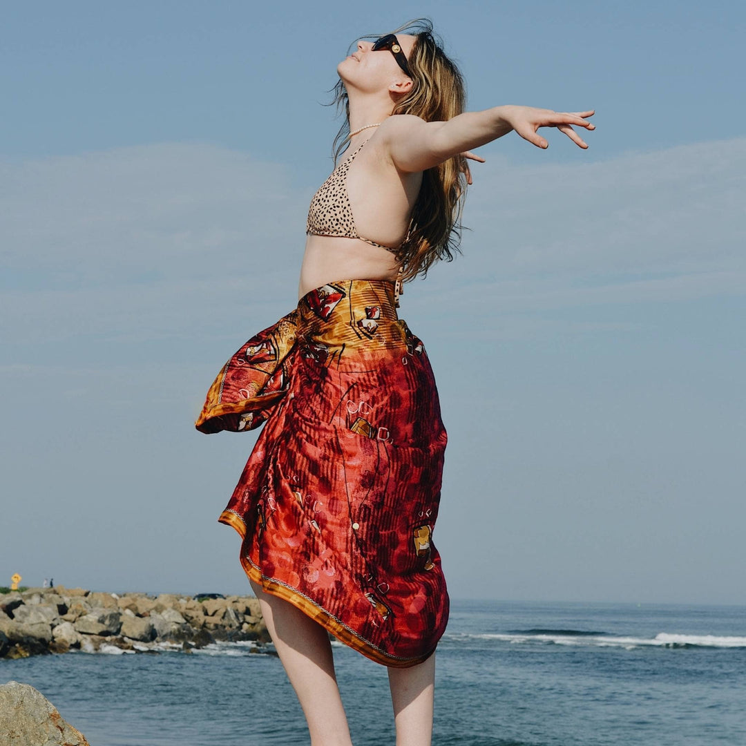 Model is wearing a red and yellow sari silk sarong scarf while standing in front of the ocean. 