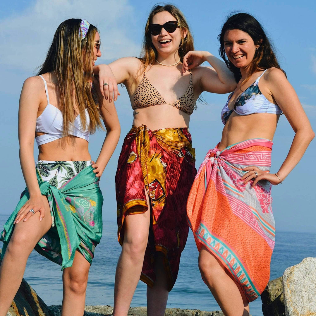 Three woman standing on a rock by the ocean wearing bikinis, covering the lower half with a sari silk beach sarong. One model is wearing a sea foam green sarong with warmer floral pattenrs, one is wearing a shimmering red sarong with gold accents. One is wearing a coral pink sarong with light blue accents.