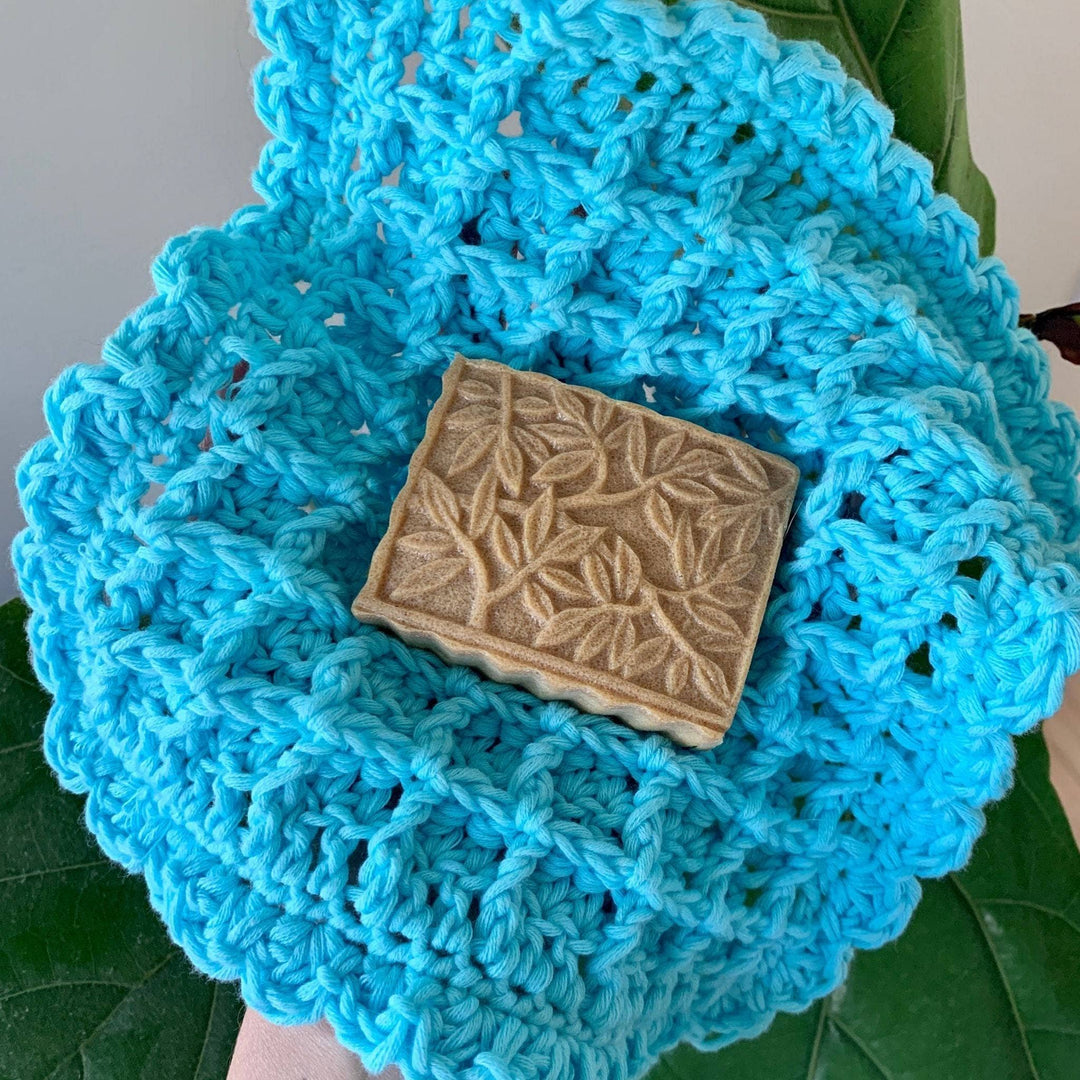person holding a bar of soap on top of blue crocheted waffle stitch wash cloth with plants in the background. 