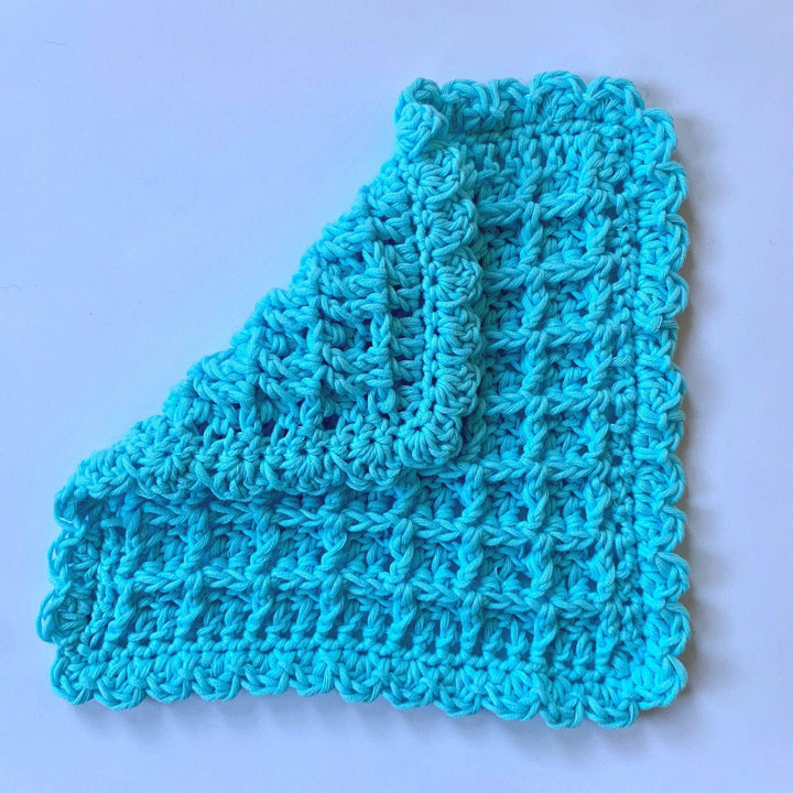blue crocheted waffle stitch washcloth partially folded over in front of a white background. 