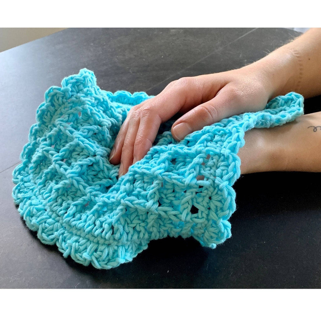hands holding a blue crocheted waffle stitch washcloth in front of a dark table.