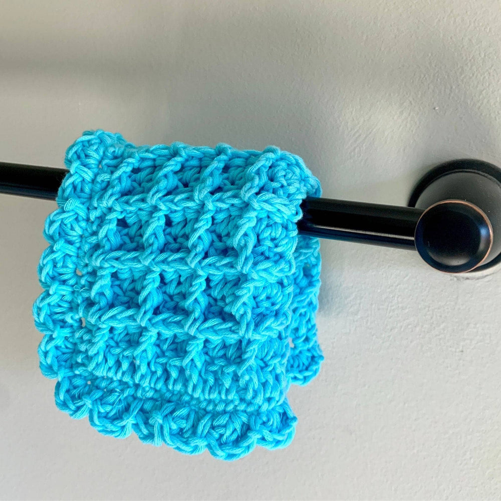 Blue crocheted washcloth hanging from dark towel bar in front of a white background. 