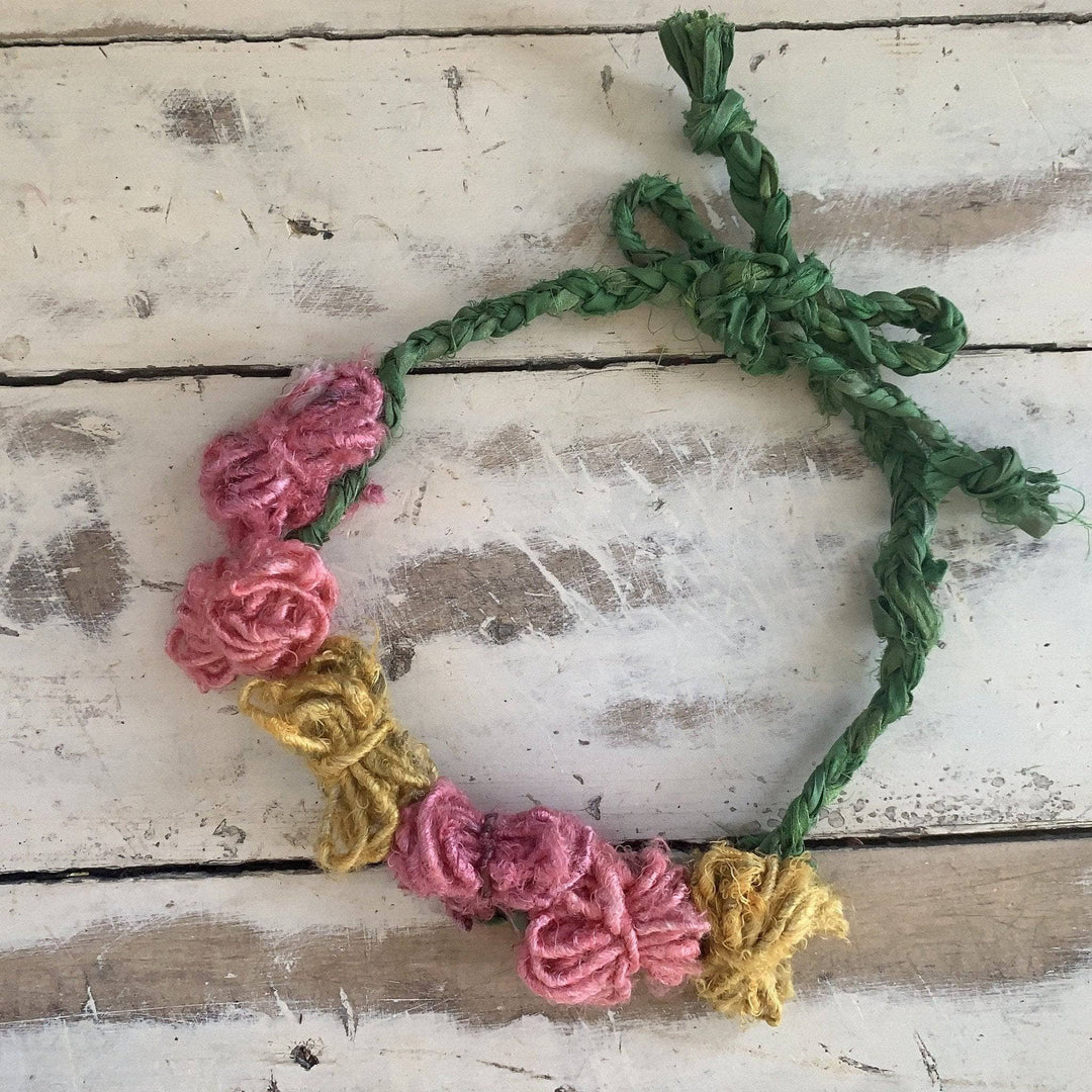 yarn flower crown (green braided band with pink and yellow "flowers") laying on a distressed wood background. 