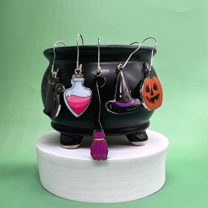 Spooky stitch marker set hanging from a black cauldron on a white pedestal in front of a green background.