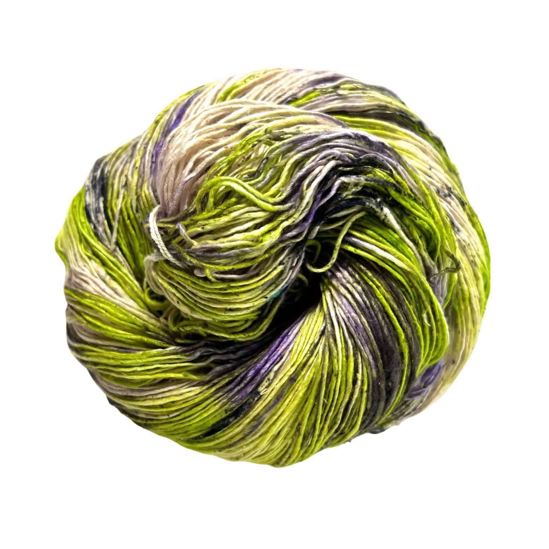 A skein of lace weight silk yarn made from reclaimed material on a white background. The skein of yarn is undyed, lime green with speckled areas of dark purple.