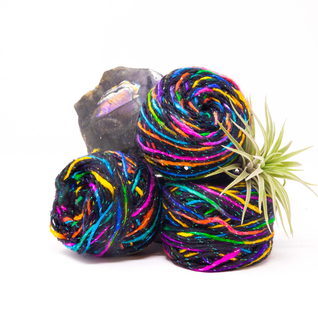 Three cakes of black and multicolored yarn with sparkle elements next to plant and stone on white background